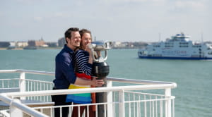Young woman looking through a viewing post aboard a Wightlink ferry is being embraced by her male partner from behind.   